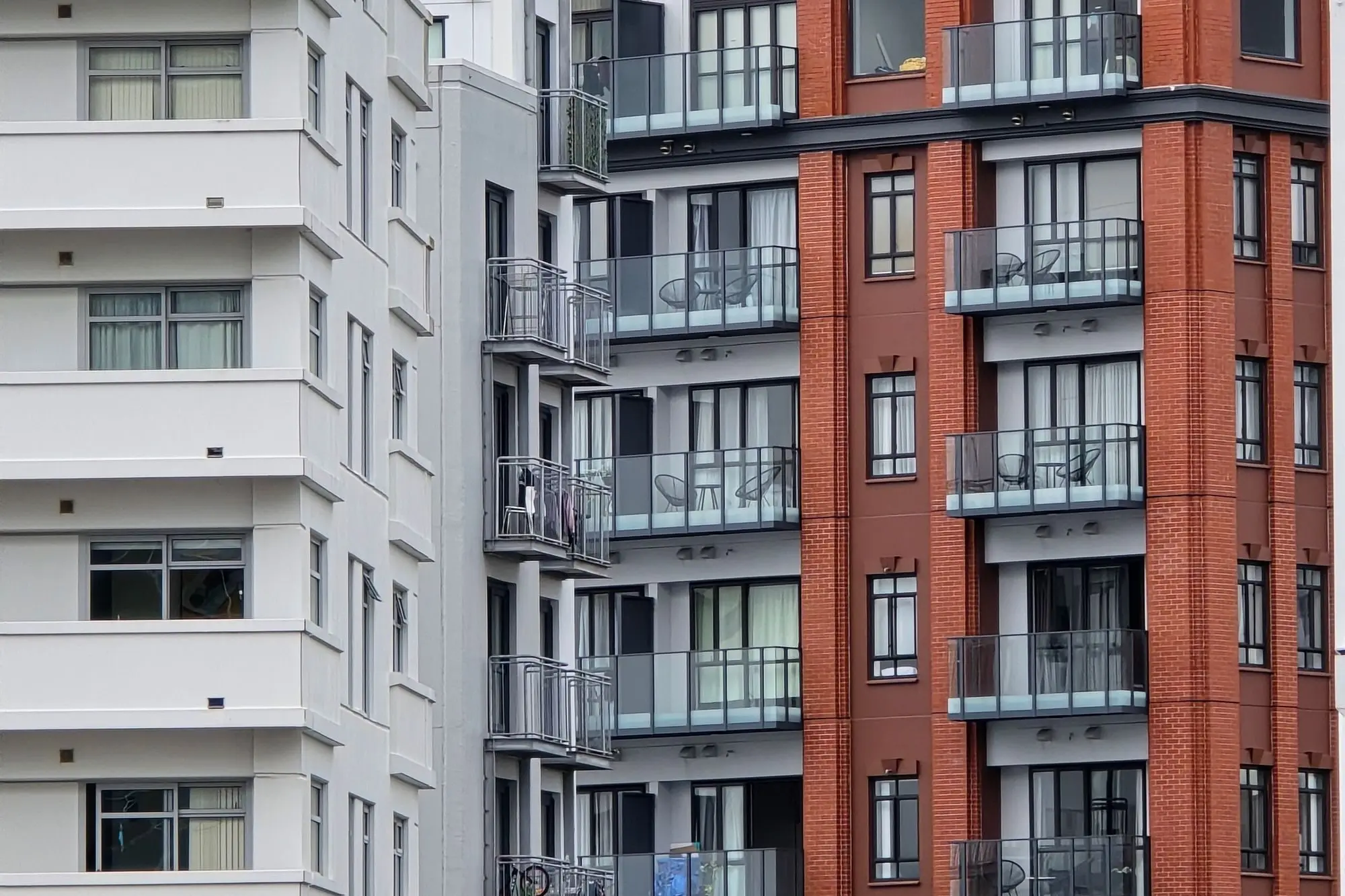 Neighbouring Auckland apartment blocks, one white and one red brick, with rows of glass balconies and outdoor chairs