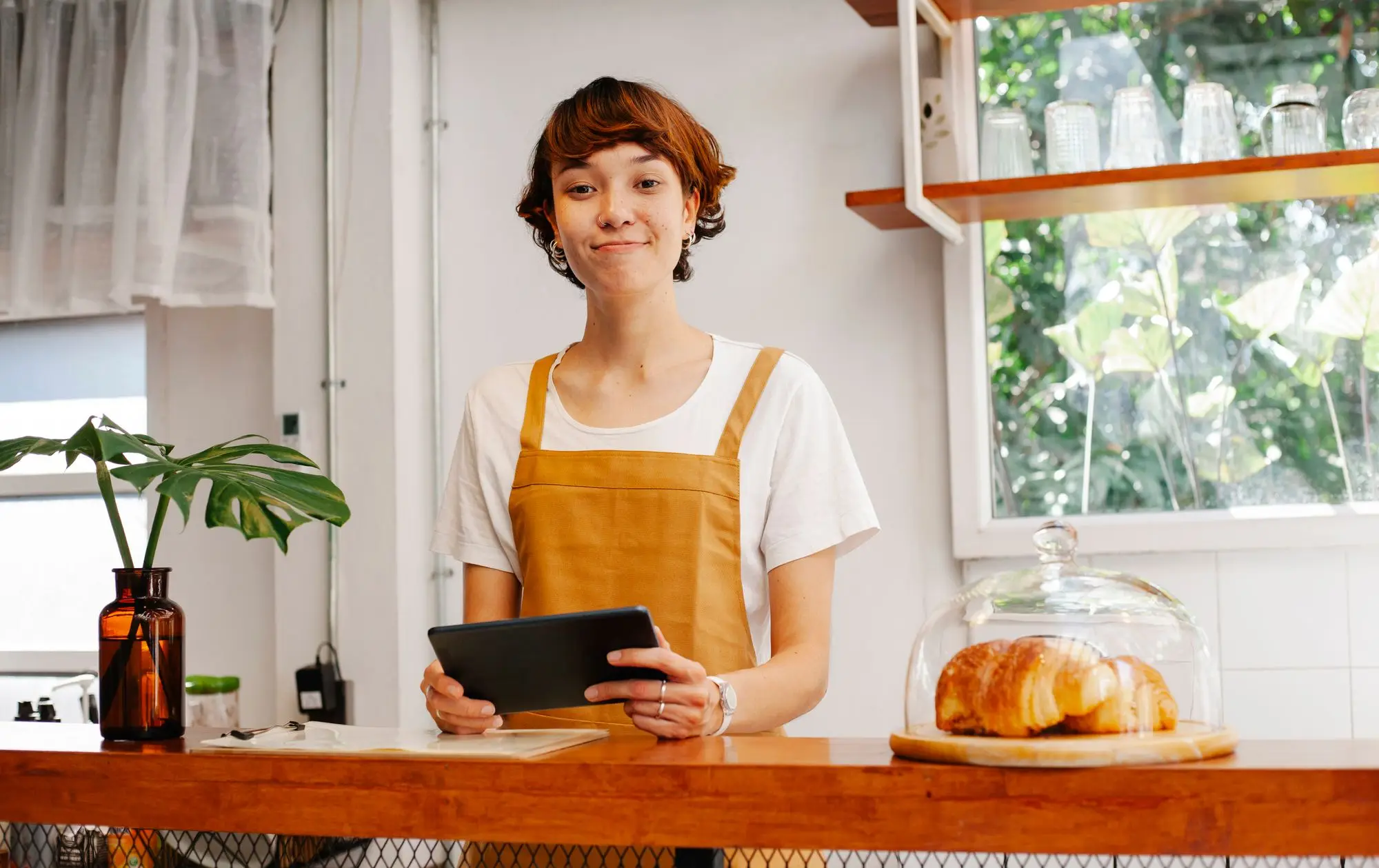 Café worker in a mustard apron holding a tablet at the counter beside a glass dome of croissants